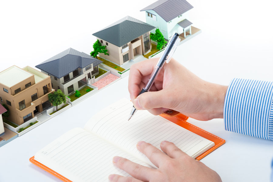 一列に並んだ住宅模型を前にして、ノートにペンで計画を書き込んでいる