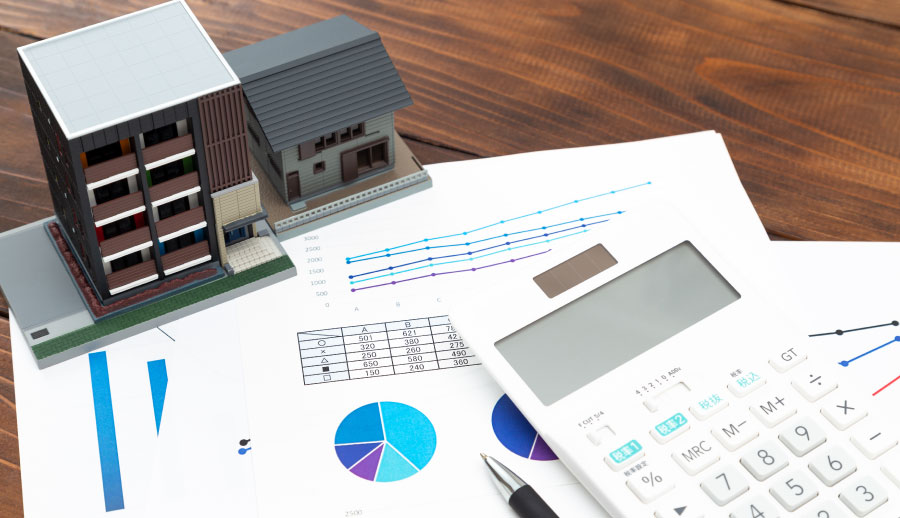 新築の家とアパートの模型、電卓、グラフや円グラフが印刷された書類、ペンが木製のテーブルに置かれている