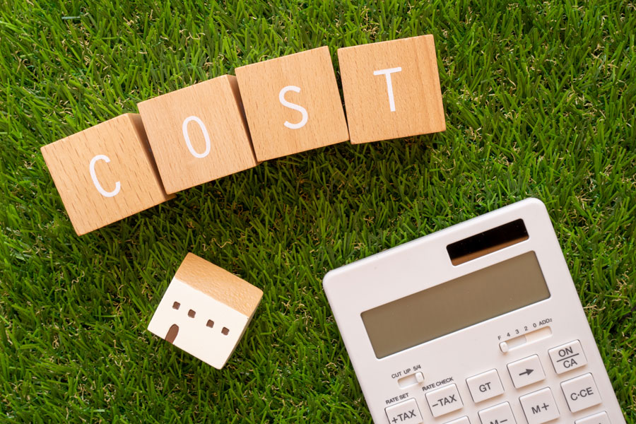 「COST」と書かれた文字ブロックと、家の模型、電卓が芝生の上に置かれている